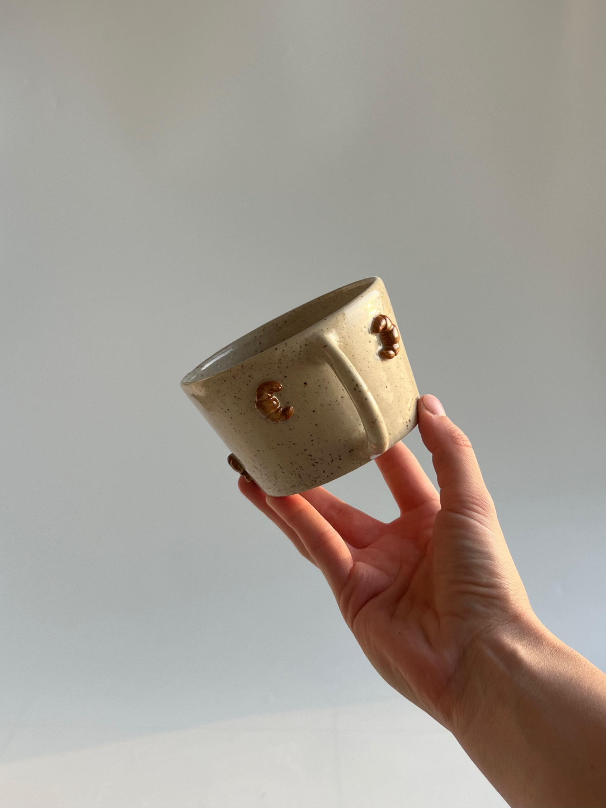 Hand holding a small beige ceramic mug with croissant decorstions, against a plain background