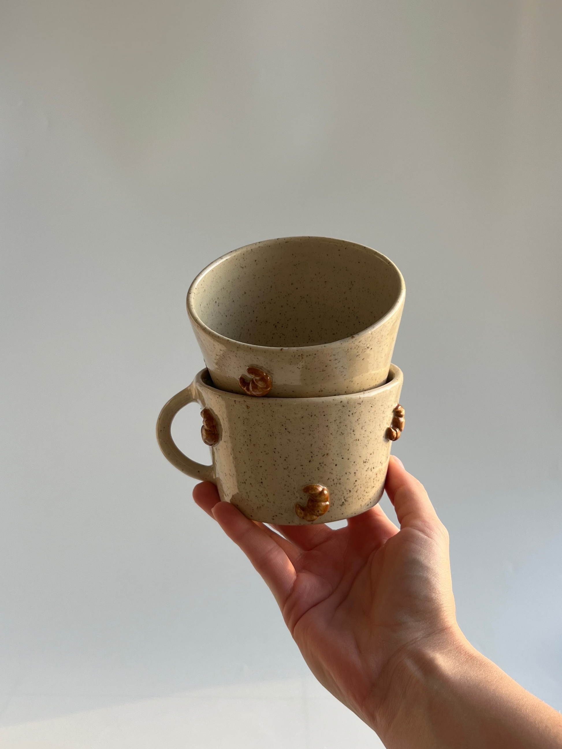 Hand holding two stacked ceramic cups with croissant decorations, against a plain background
